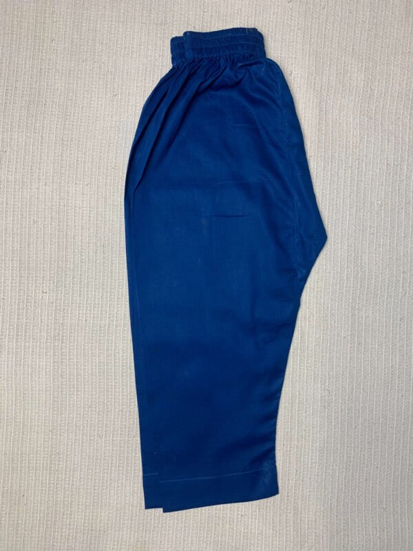 A pair of deep blue, cropped pants with an elasticated waistband, laid flat on a light-colored, textured surface. The pants appear to be made of a solid, matte fabric. The legs are straight and cut off around the mid-calf.